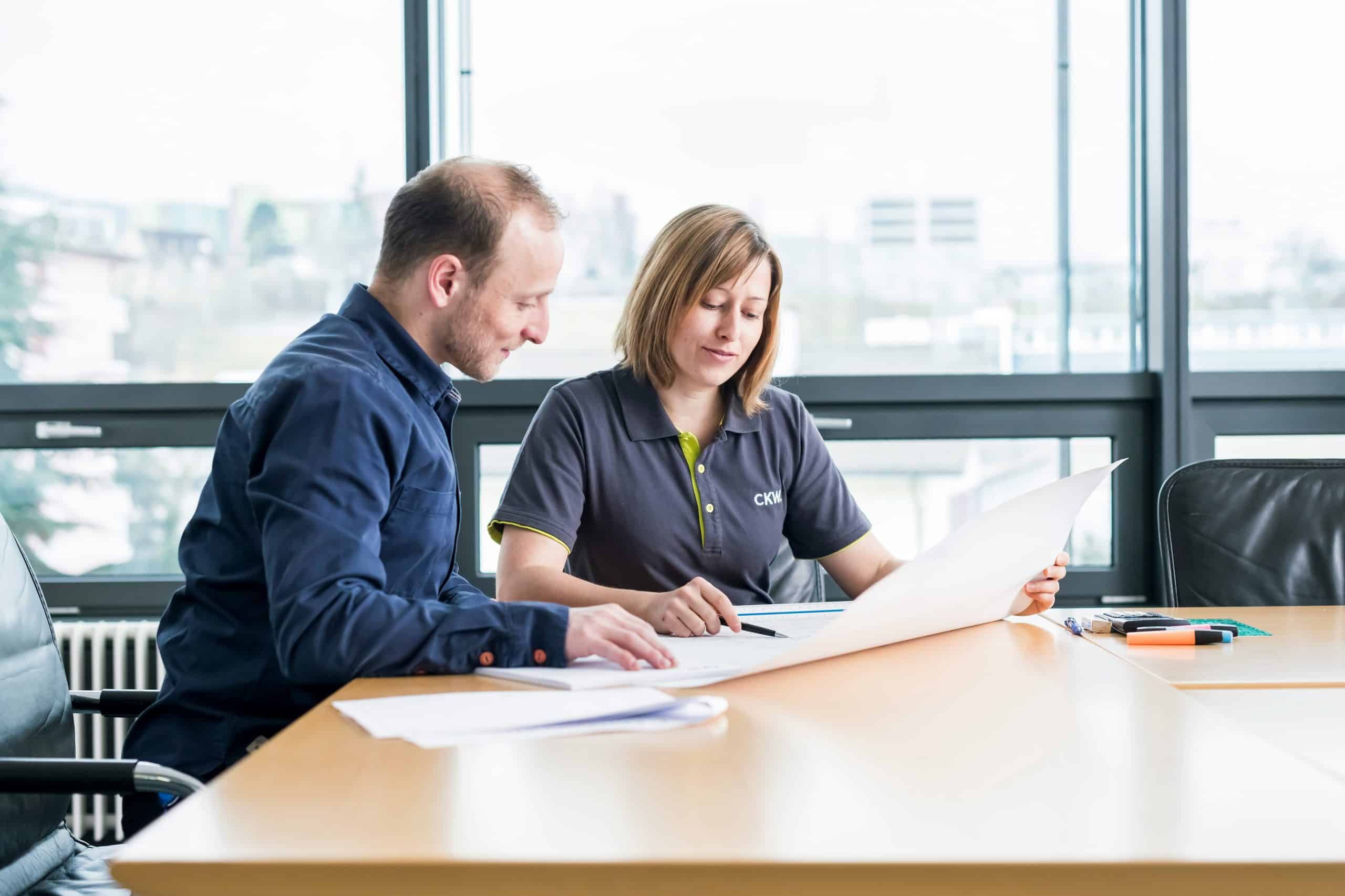Two people sit at a conference table in a bright office, reviewing large documents together. One person is wearing a dark shirt, the other a polo shirt. Papers, pens, and a marker are on the table.