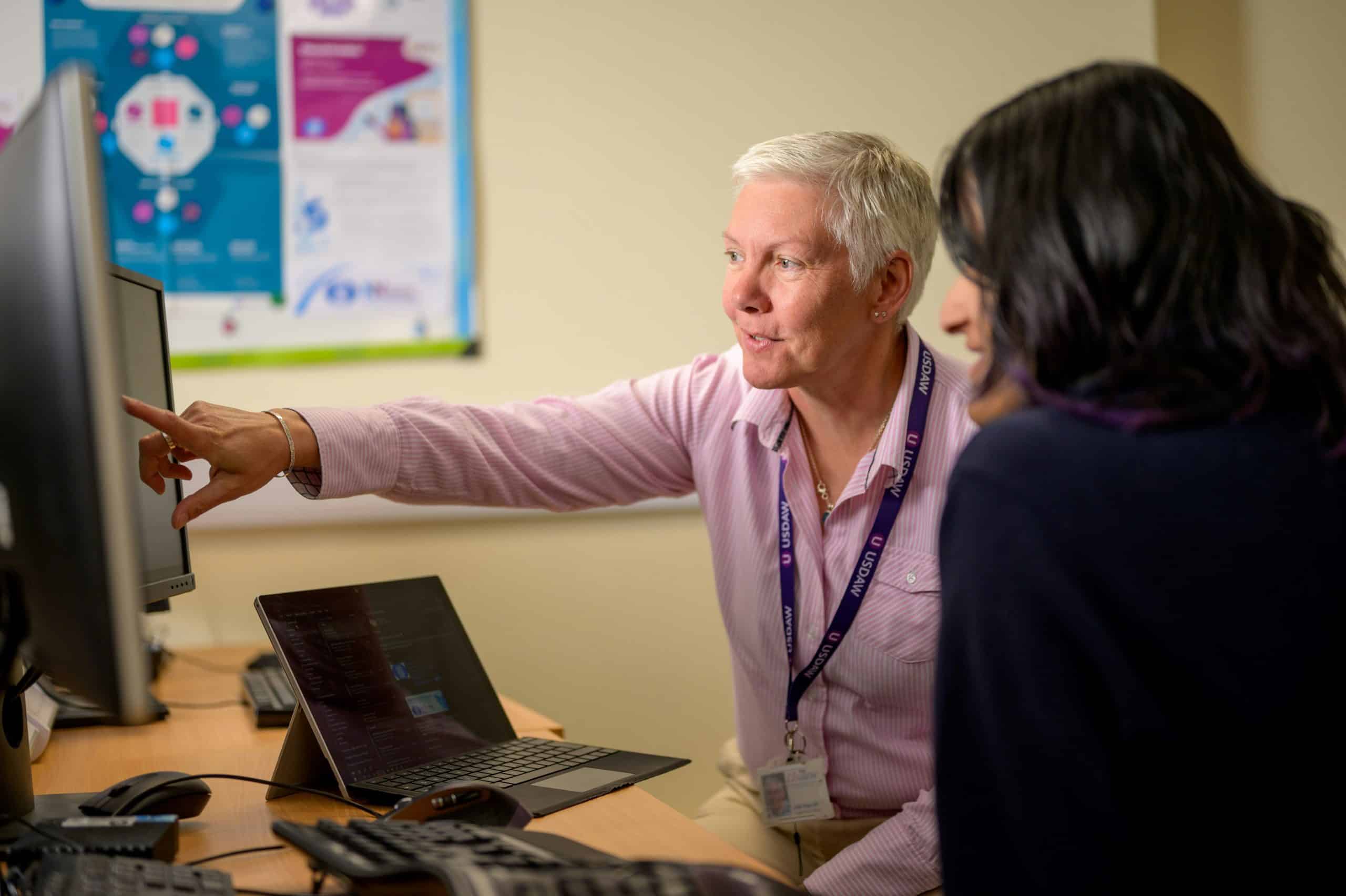 Two people sit at a desk with computers. One person, wearing a lanyard, points at the monitor while explaining something to the other, who listens attentively. A colorful poster is visible on the wall behind them.