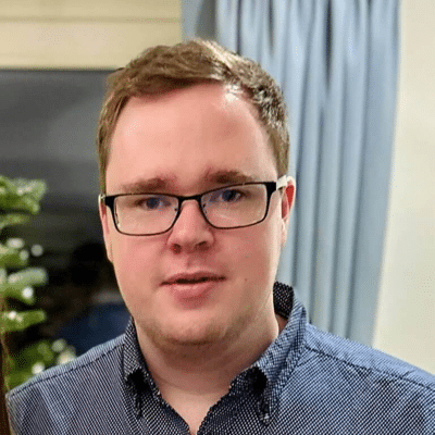 A young man with short light brown hair and glasses, representing About Techwise Support, is wearing a navy blue button-up shirt and stands indoors in front of a window with blue curtains.