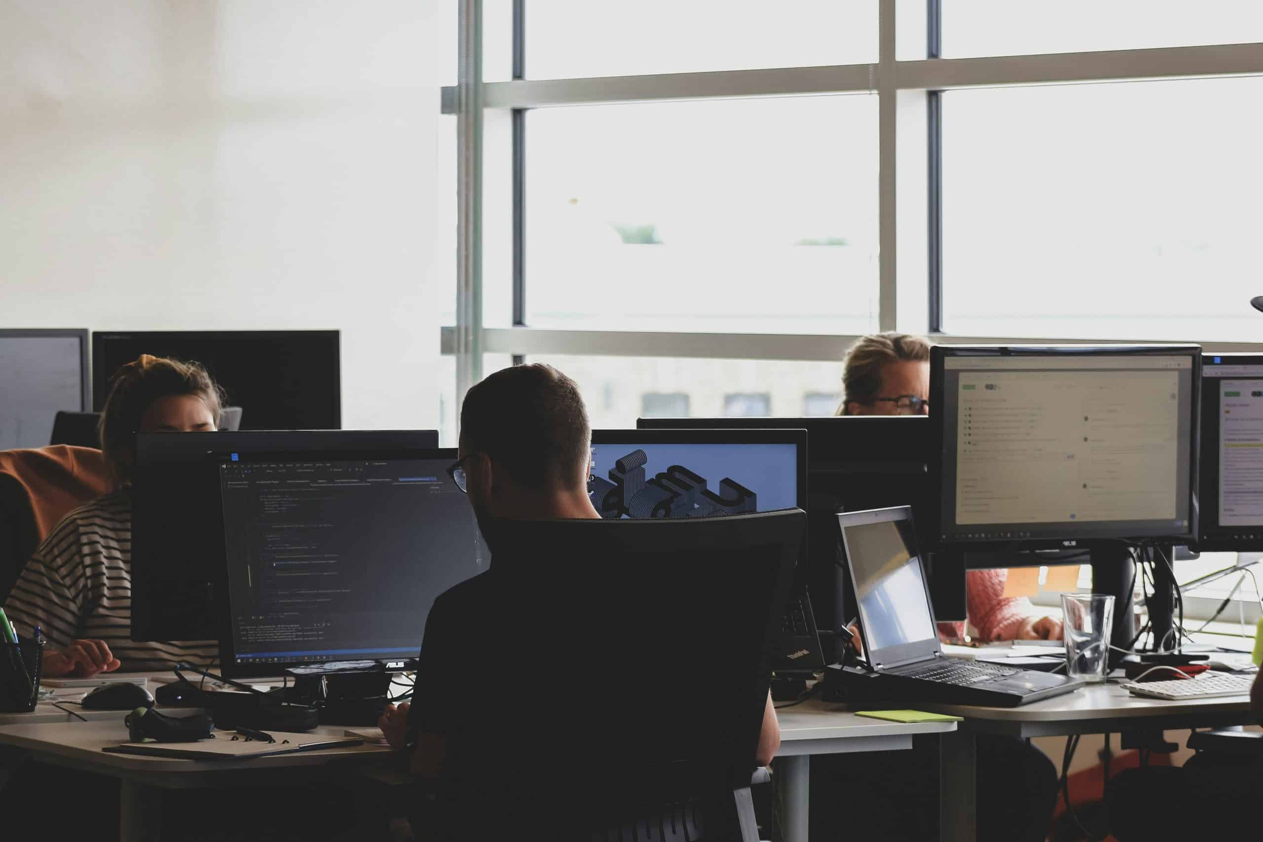 Three people work at desks with multiple computer monitors in a modern, brightly lit office, showcasing a collaborative environment ideal for Business IT Support & Managed IT Services. Large windows provide plenty of natural light in the background.