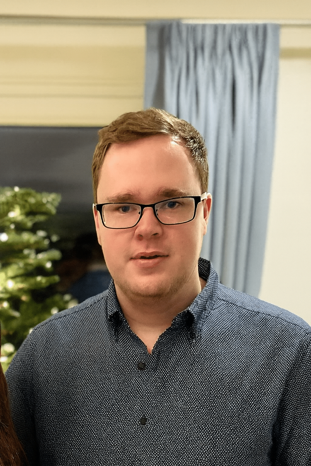 A young man with short brown hair and glasses, wearing a dark blue button-up shirt, stands indoors in front of a curtain and a lit Christmas tree.