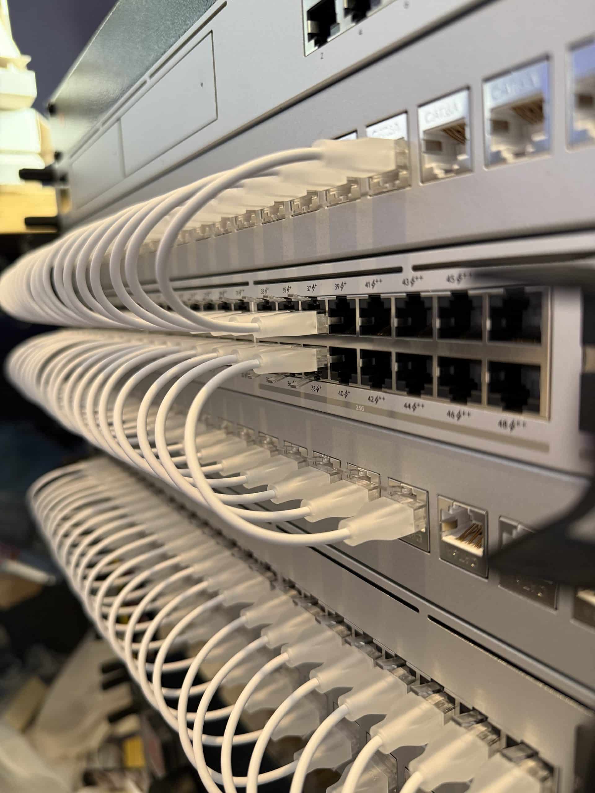 Close-up of multiple white Ethernet cables neatly plugged into several rows of network switches in a server rack, showcasing organized cable management and robust Unifi Networking and Security Solutions infrastructure.