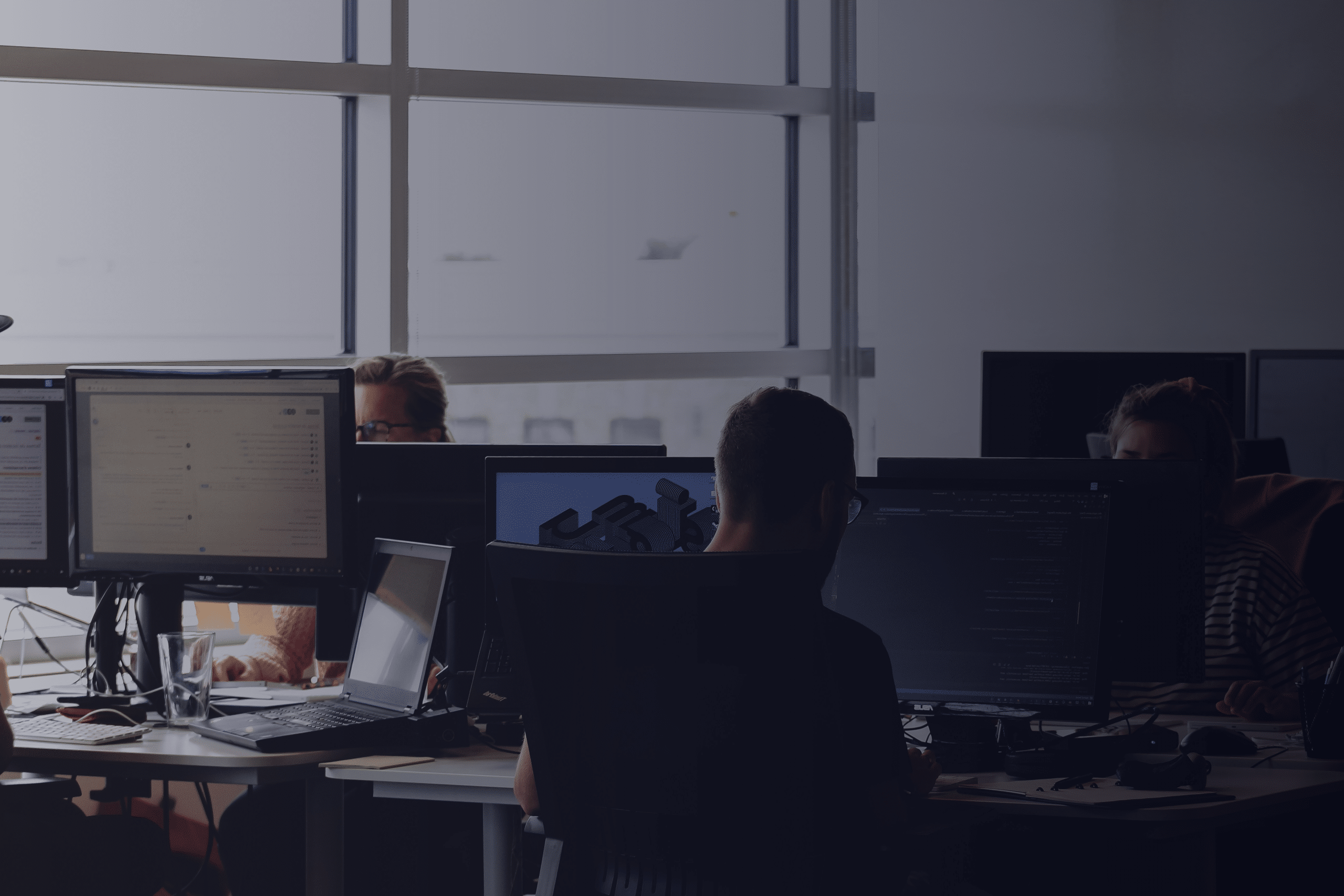 Several people work at desks with multiple computer monitors in a modern, dimly lit office. Sunlight filters through large windows in the background, illuminating parts of the Techwise Support workspace.