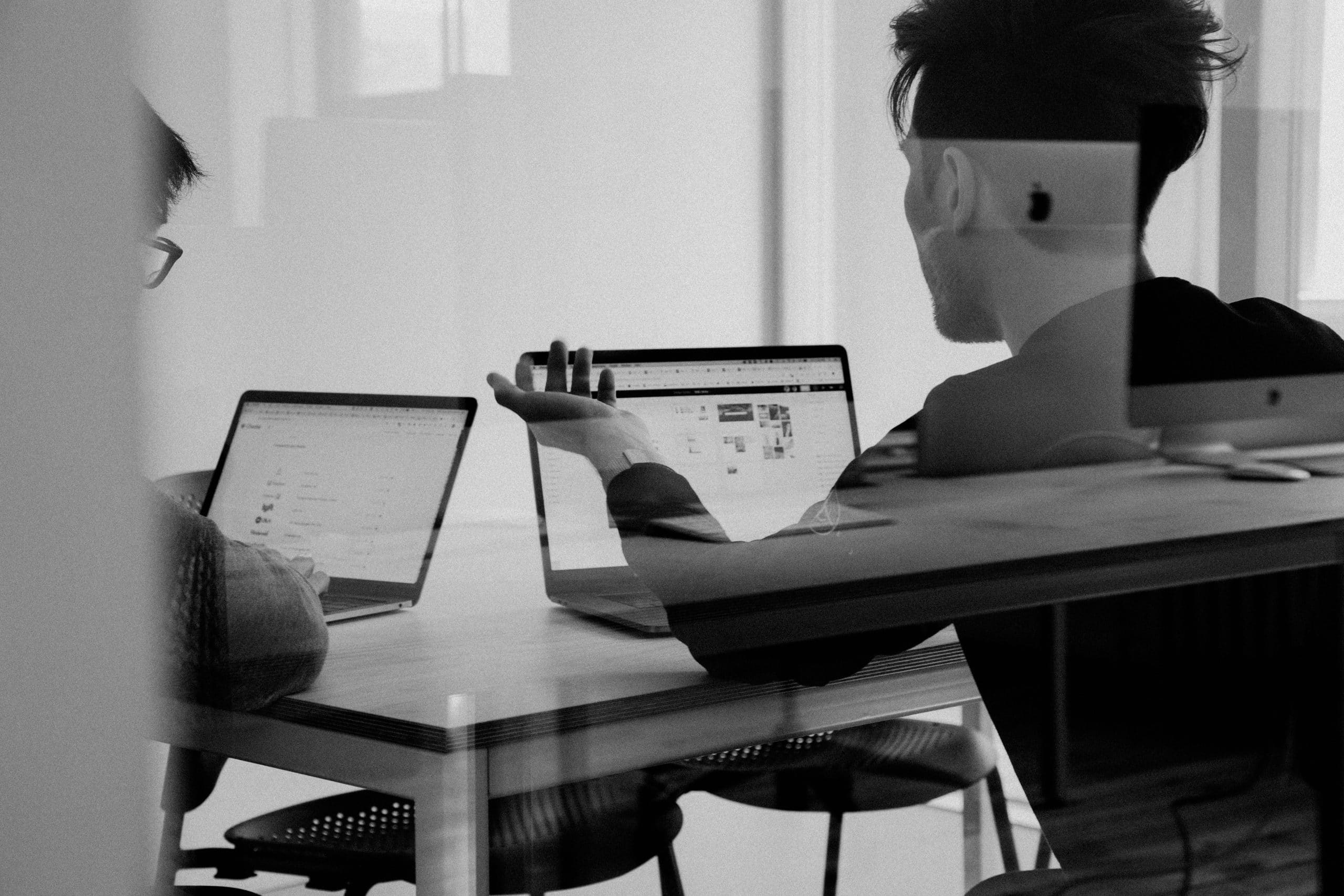 Two people sit at a table with open laptops, discussing something on the screens. The black-and-white photo, taken through glass, captures reflections and a collaborative office atmosphere focused on technology and start-up IT support.