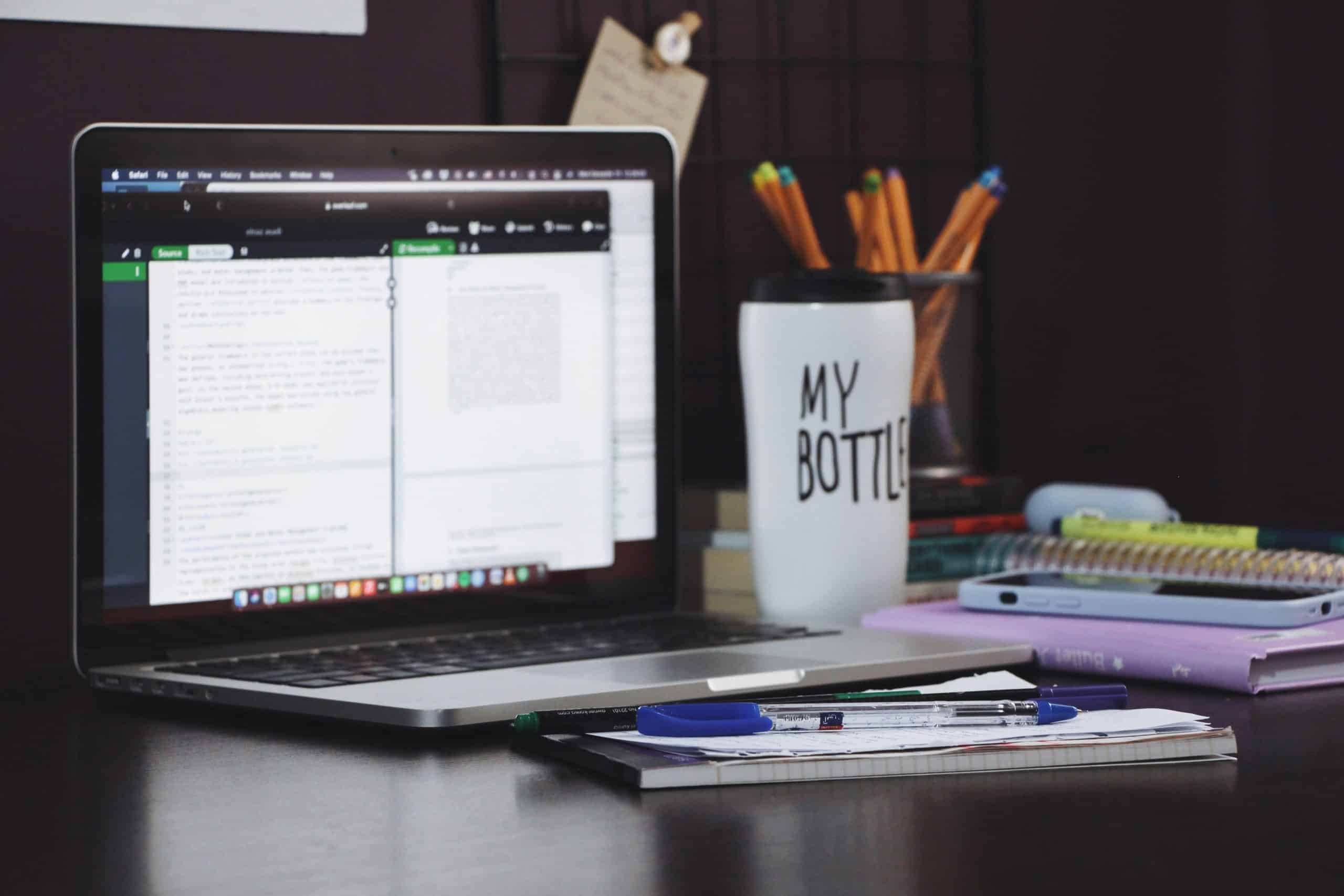 A laptop displaying code sits on a desk next to a notebook with pens, a reusable cup labeled MY BOTTLE, a calculator, and some colored pencils in a holder. Books and notepads are stacked in the background.