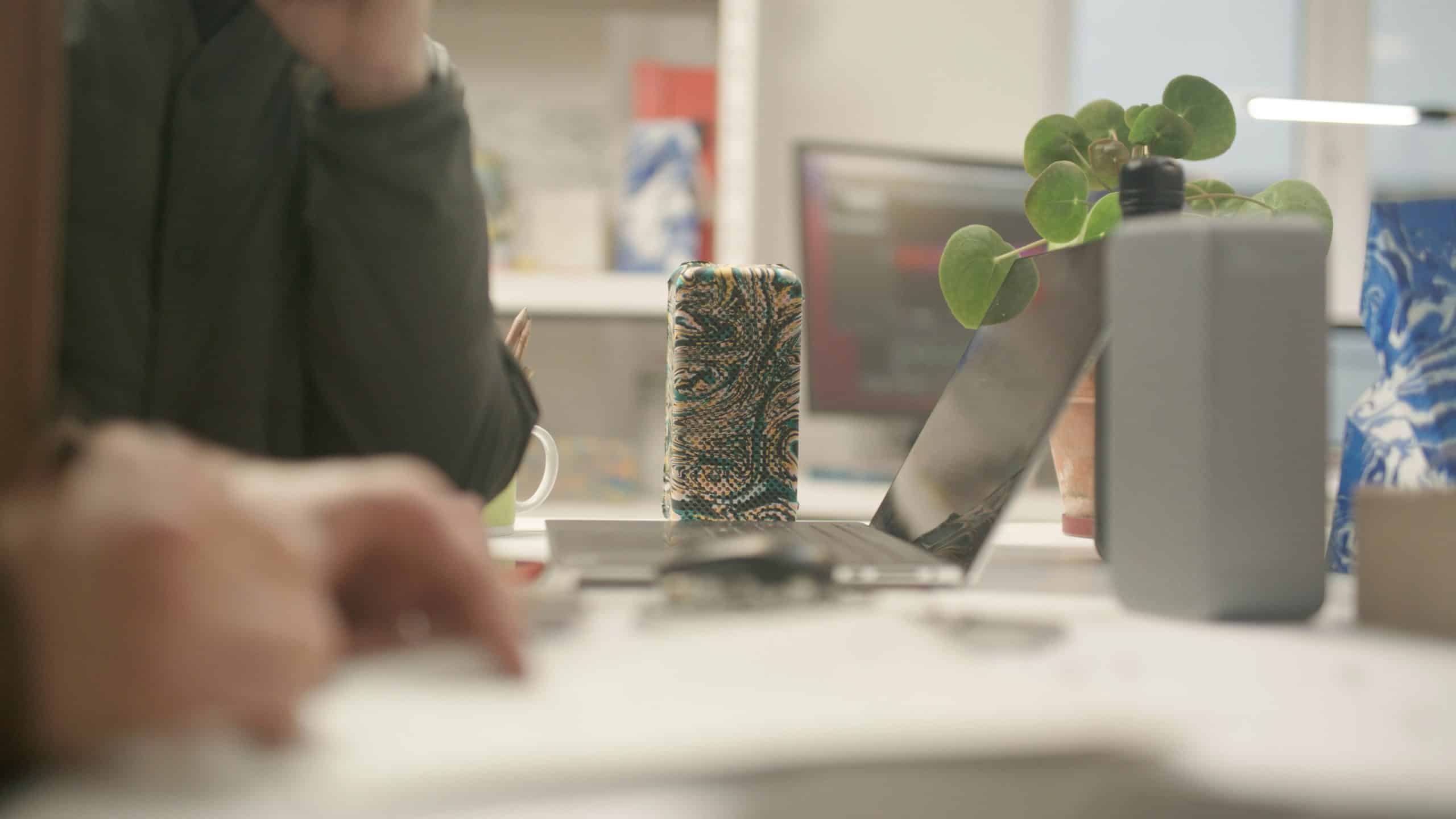 A close-up of a cluttered desk with papers, a laptop, a plant, and a patterned cup. A person is blurred in the background, partially visible and seated at the desk.