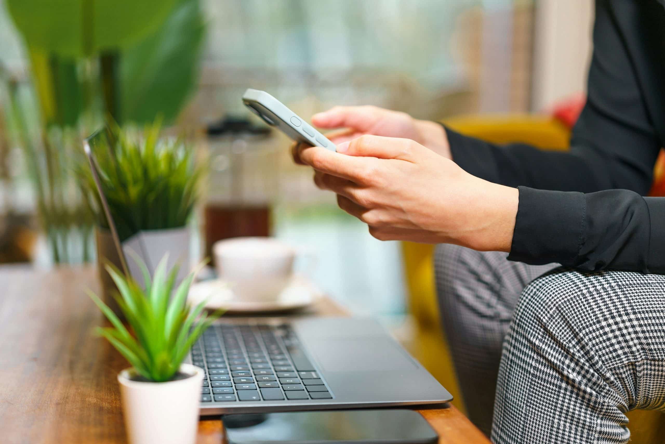 Person sitting at a table using a smartphone with an open laptop in front. A cup of coffee, two small potted plants, and a smartphone are also on the wooden table. The background is softly blurred.