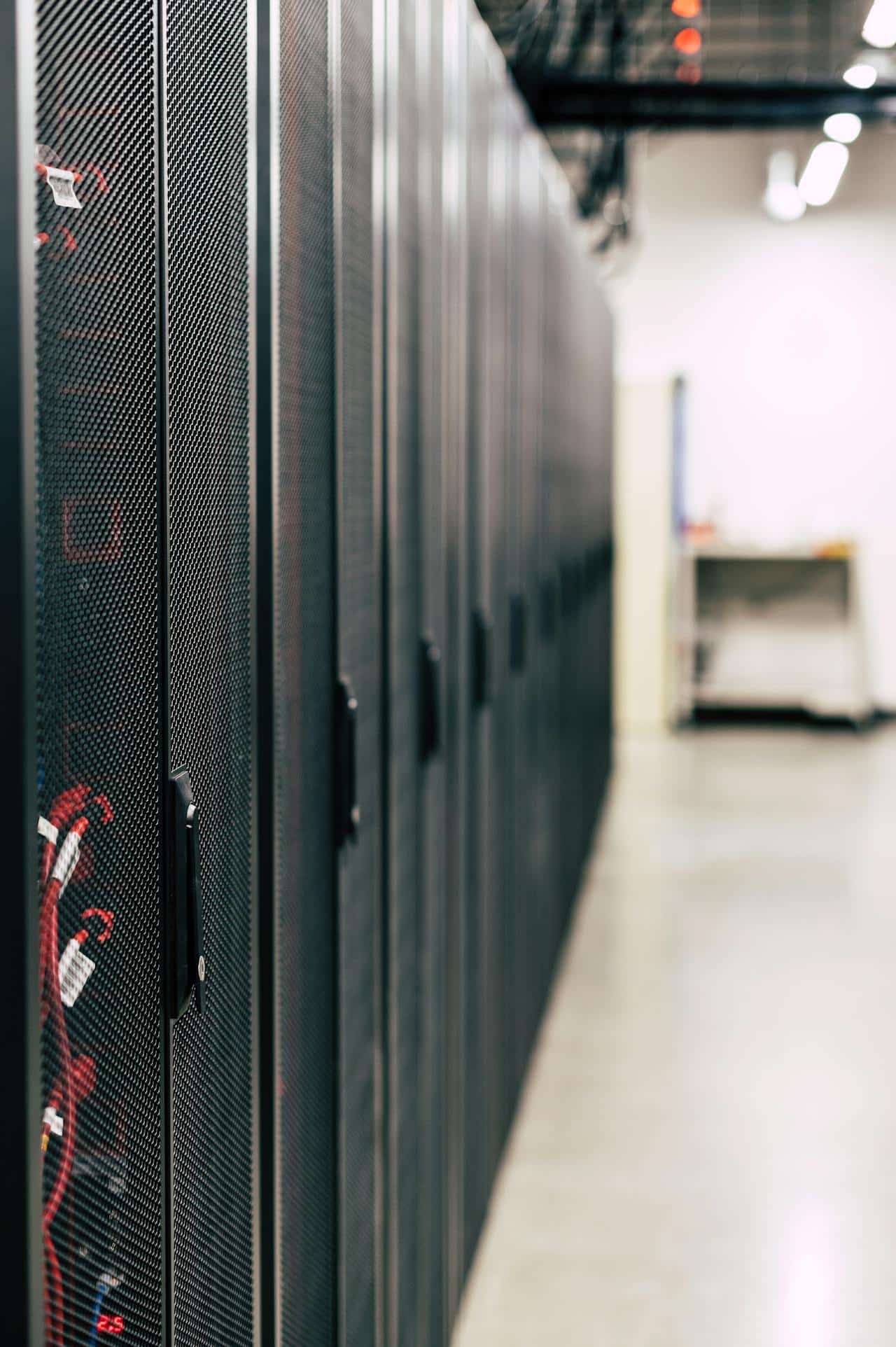 A row of black server racks with mesh doors stands in a brightly lit data center at a Datto IT Solutions Partner site, with cables visible inside one rack and blurred office equipment in the background.