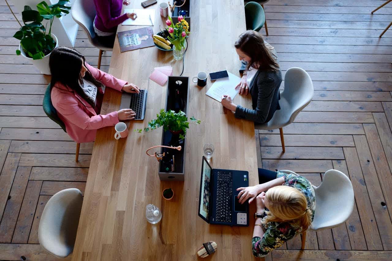 Three women work at a long wooden table with laptops, notebooks, and coffee cups. The workspace, ideal for IT support for business productivity, is decorated with plants, flowers, natural light, and a wooden floor. The view is from above.