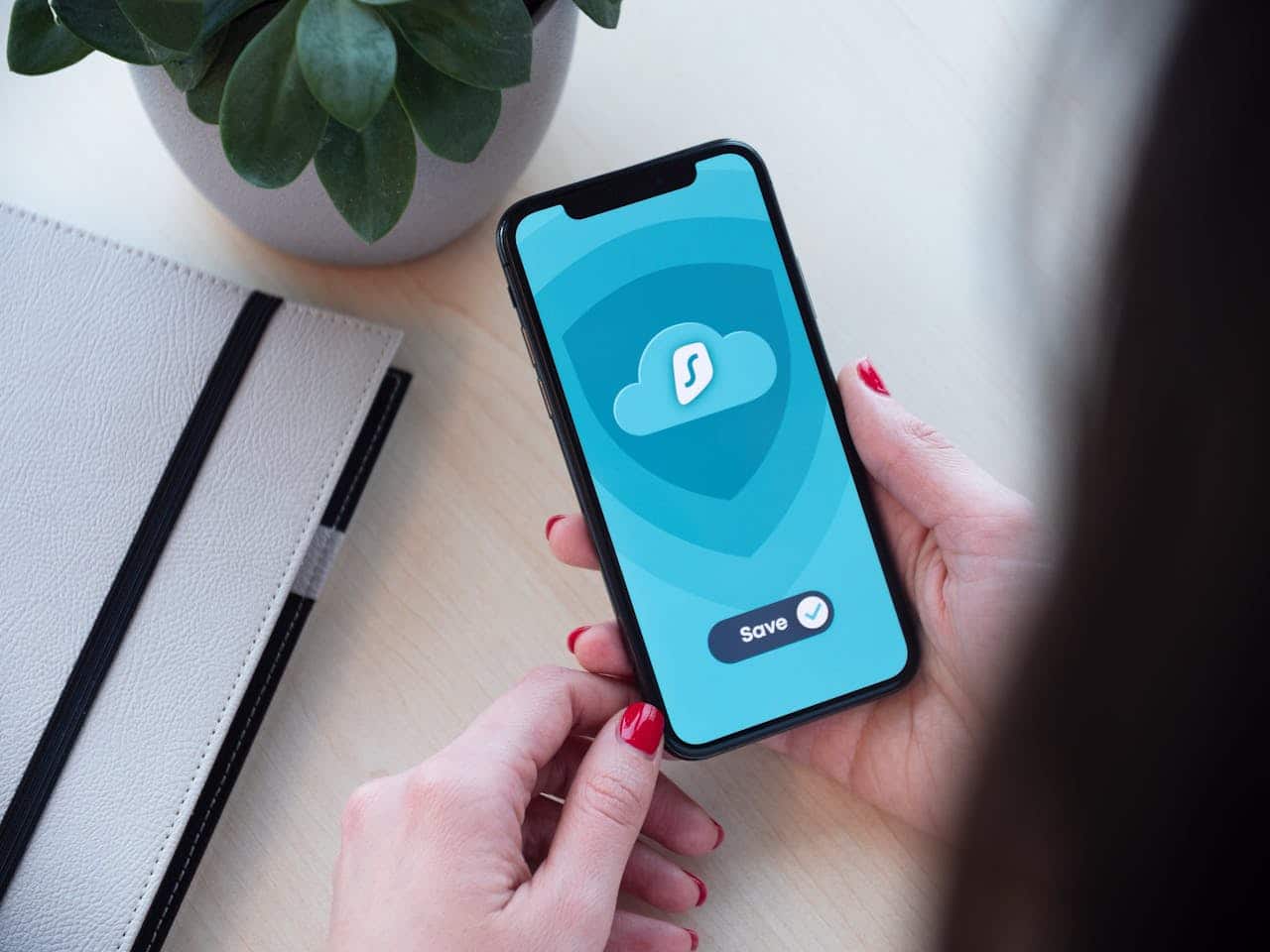 A person with red nail polish holds a smartphone displaying a blue screen with a cloud icon and a Save button—perfect for accessing Top Tips For Home Office Security. A notebook and potted plant rest on the light-colored desk.