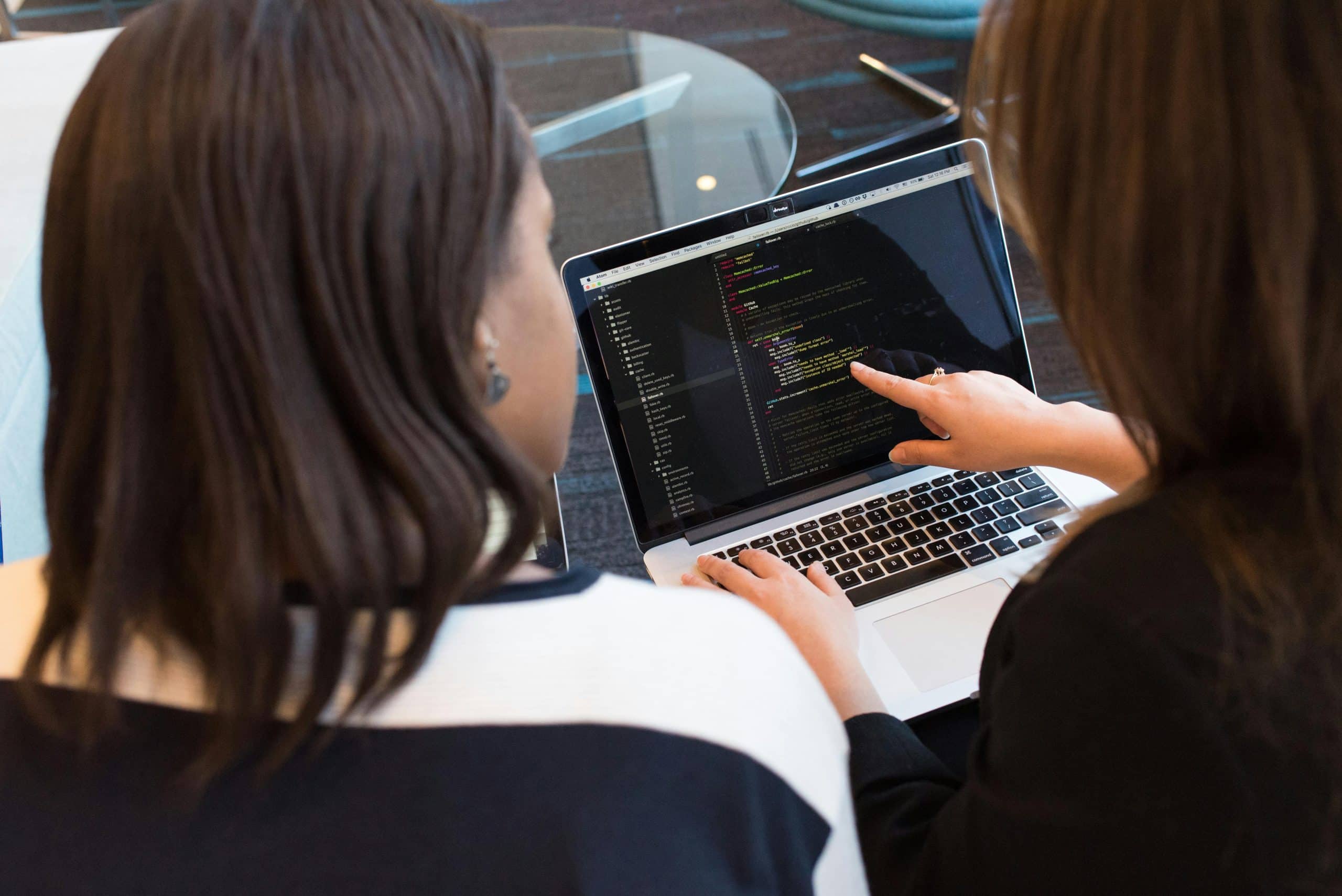 Two women sit side by side at a table, looking at a laptop screen displaying lines of code. One woman points at the code while the other observes, illustrating a collaborative IT consultancy or programming session.