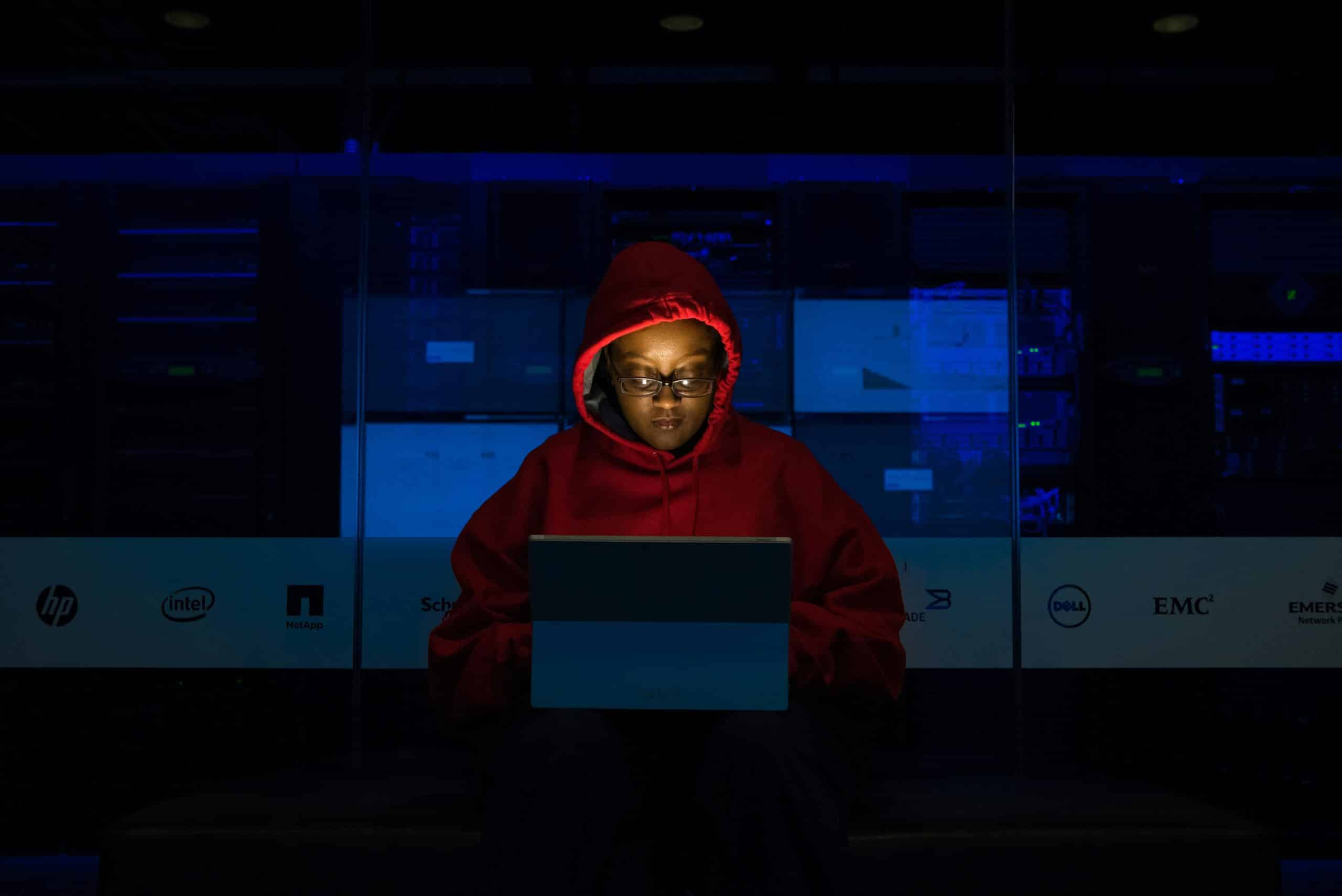 A person wearing a red hoodie sits in a dark room, illuminated by the light from a laptop screen. Computer servers and Security & Compliance Solutions logos are visible in the background.