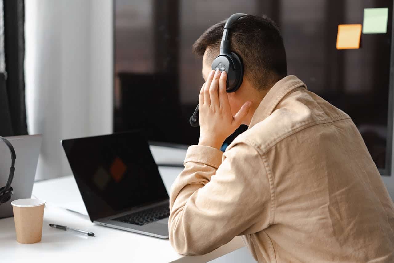 A person wearing headphones sits at a desk, facing a laptop with a hand resting on their face—perhaps awaiting an answer from an IT support company. A paper cup, pen, and another laptop sit on the white desk; sticky notes cover a black monitor in the background.