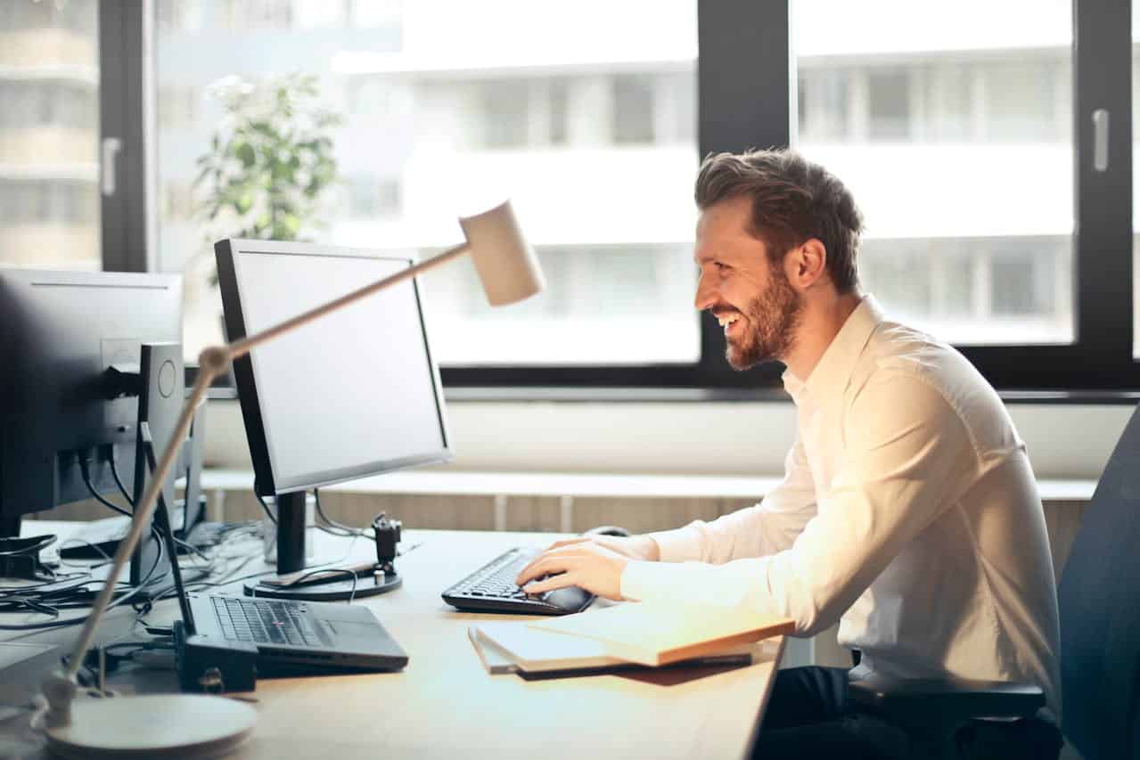 A smiling man in a white shirt provides IT support for business productivity at his desk with two monitors, a lamp, a notebook, and office supplies in a bright office with large windows.