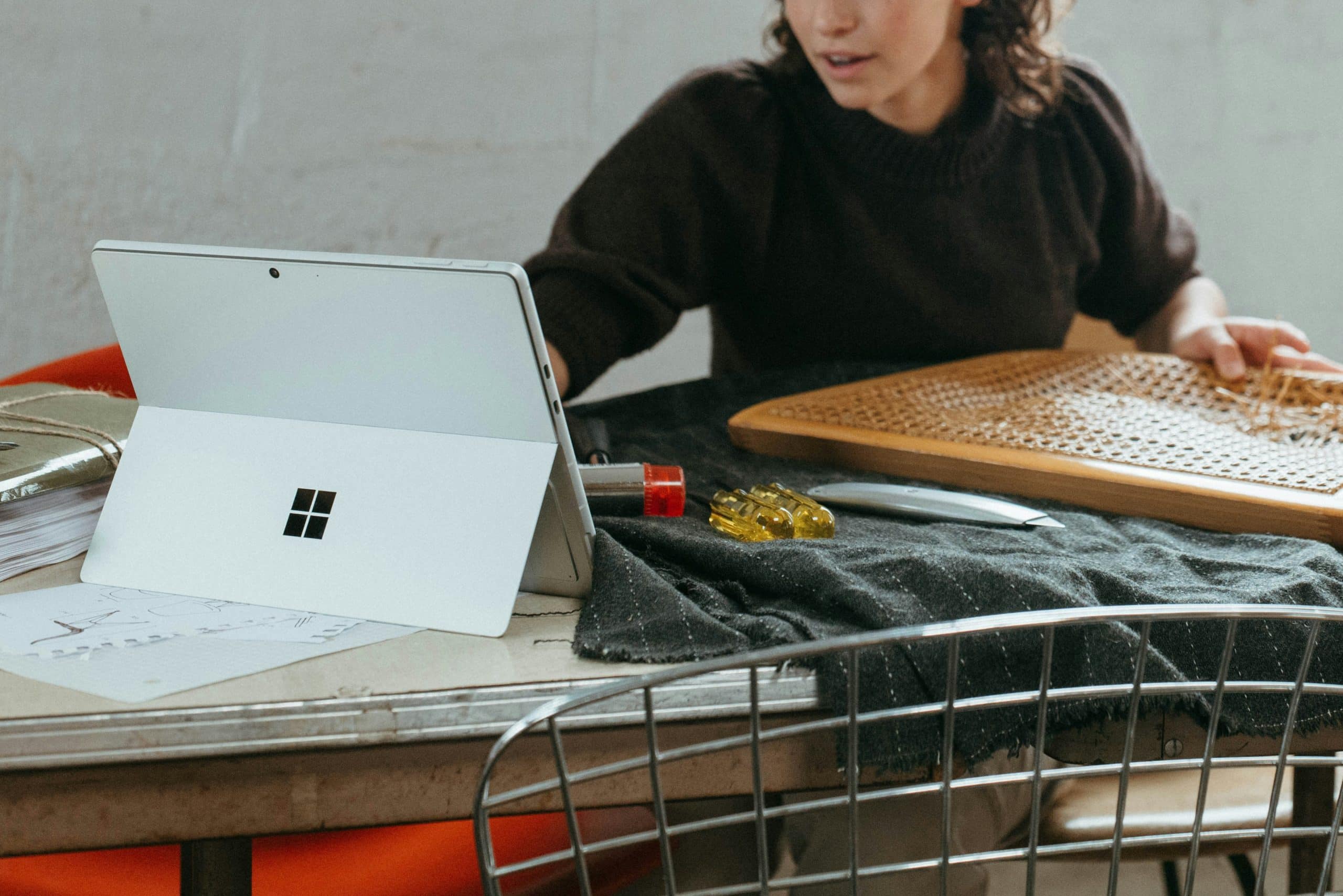 A woman sits at a table with a Microsoft Surface tablet, papers, a pen, and a knitting project, partially out of view. She appears to be working or studying while crafting.
