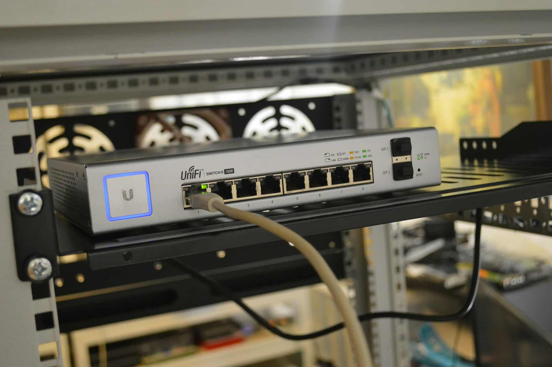 A UniFi network switch from UniFi Networking and Security Solutions sits on a rack shelf, with multiple Ethernet cables plugged into its ports. The device is powered on, indicated by lit LED lights. Computer equipment and cables are visible in the blurred background.